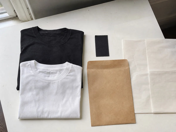 Photorealistic editorial image of a minimal clothing-line setup with two blank T-shirts, a swatch and plain packaging.