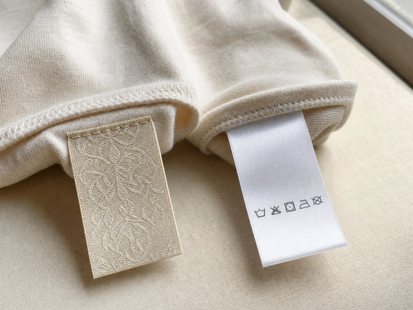 Photorealistic close-up of a woven garment label and a printed satin care label sewn into cotton fabric.