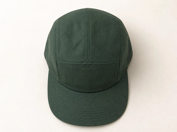 Photorealistic product mockup of a blank forest green cotton twill five-panel cap viewed overhead on an off-white background.