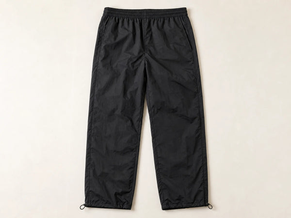 Photorealistic product mockup of blank black matte crinkle-nylon straight-leg track pants with adjustable hems viewed overhead.