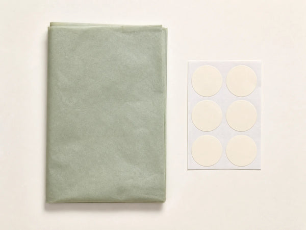 Photorealistic product mockup of folded sage garment tissue beside six blank cream circular sealing stickers.