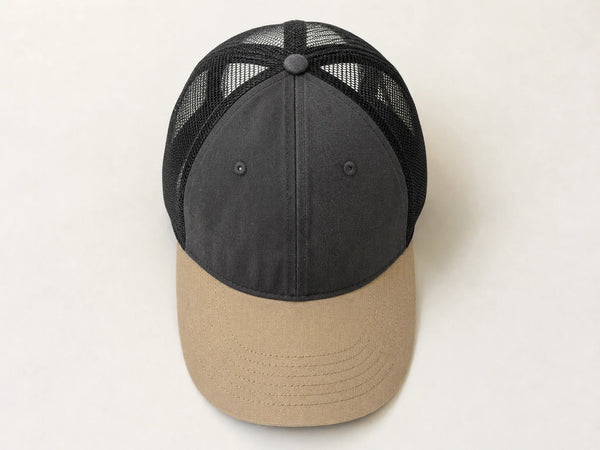 Photorealistic product mockup of a blank charcoal mesh trucker cap with a tan curved brim viewed overhead on an off-white background.